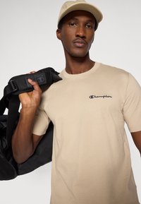 Μπεζ κοντομάνικη μπλούζα με κεντημένο λογότυπο "Champion", κρατούμενη από ένα άτομο που έχει μαύρη τσάντα duffel. Το καπέλο είναι ανοιχτό μπεζ.