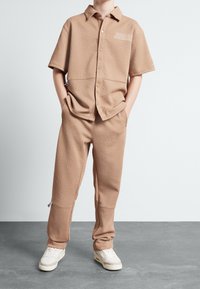 Tan t-shirt med kort ärm och matchande byxor med ett strukturerat geometriskt mönster. Innehåller en subtil logotyp och parats ihop med ljusa sneakers.