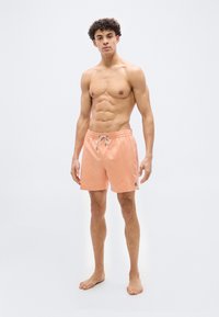 Ευθυτενής νεαρός άνδρας που στέκεται ξυπόλυτος φορώντας ανοιχτό πορτοκαλί μαγιό με κορδόνι, μπροστά σε απλό λευκό φόντο.