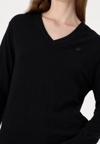 Pull-over noir en tricot avec décolleté en V, doté de manches longues et d'un logo subtil sur la poitrine. Texture douce et design ajusté.