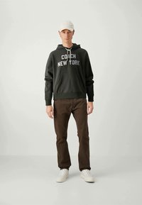 Sötétzöld kapucnis pulóver, amelyen fehérrel van nyomtatva a "COACH NEW YORK" felirat, barna nadrág és fehér sportcipő. A világos színű sapka teszi teljessé a megjelenést.