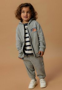 Felpa grigia con zip e tasche frontali, con logo arancione "Stadium". Abbinata a pantaloni della tuta grigi e scarpe bianche. Camicia a righe sotto.