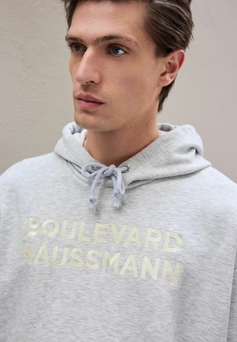 Grey hoodie with drawstring, featuring metallic text "BOULEVARD HAUSSMANN" on the front. Soft texture, casual design, relaxed fit.