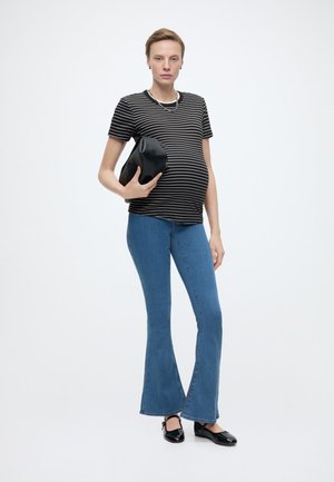 Grūtniece, valkājot melnu un baltu svītrainu krekla, zilas platām biksēm, melnus apavus, turot melnu somiņu, stāv uz balta fona.