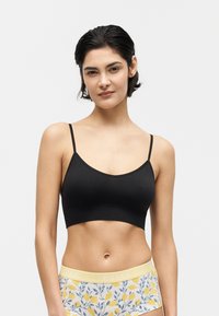 Vrouw met kort donker haar die een zwart cropped hemdje en hoog getailleerd bloemenondergoed met citroenenprint op een lichte achtergrond draagt.