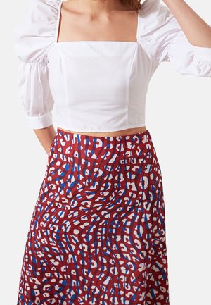 Kobieta w białym crop topie z bufkami i kwadratowym dekoltem, w połączeniu z czerwonym, wysokim stanem spódnicą z niebiesko-białym abstrakcyjnym wzorem.