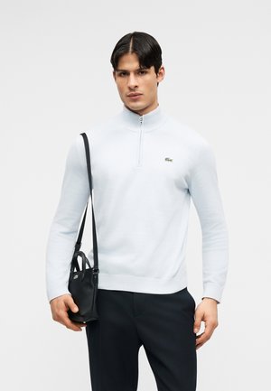 Mees valges kvartstõmblusega kampsunis ja mustades pükstes hoiab musta nahast õlakotti pika rihmaga valge tausta ees.