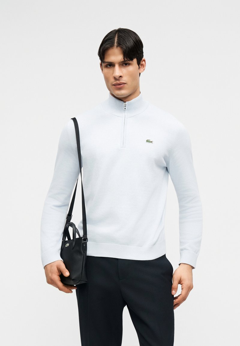 Άνδρας με λευκό πουλόβερ με φερμουάρ στο καρφί και μαύρο παντελόνι κρατάει μαύρη δερμάτινη τσάντα χιαστί με μακριά λουρί μπροστά σε λευκό φόντο.