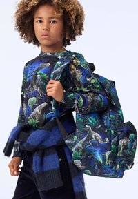Svart ryggsekk med dinosaur- og jungelmønster, med justerbare stropper. Barnet har på seg en matchende langermet skjorte med lignende design.