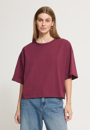 Kvinde med langt, glat brunt hår bærer en løs bordeaux T-shirt og blå jeans, står og vender fremad mod en ensfarvet lys baggrund.
