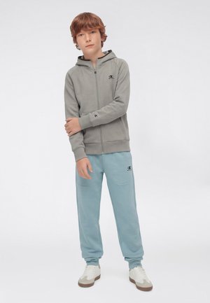 Młoda osoba stojąca w jasnoszarej bluzie na zamek, jasnoniebieskich spodniach dresowych i białych tenisówkach na białym tle.