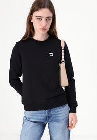 Felpa nera con scollo rotondo e logo ricamato. Abbinata a jeans azzurri chiari e una borsa a tracolla beige con la tracolla etichettata "Hugo."