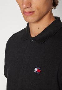 Camiseta polo negra con tejido texturizado, cuello clásico, abertura de dos botones y un pequeño logo de Tommy Jeans en el pecho.