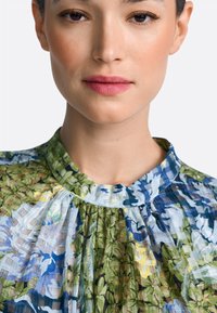 Bluză cu model floral în nuanțe de verde, albastru și galben; material ușor, plisat, cu decolteu rotund și detalii contrastante de textură.