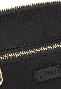 Primer plano de una bolsa de tela negra con una cremallera horizontal dorada y una etiqueta negra cosida con el nombre de la marca "TOUS" en relieve.