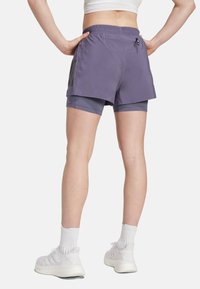 Lila sportshorts med elastisk midjeband, som har en dold ficka och grå innertyg, bärs med vita sneakers och strumpor.