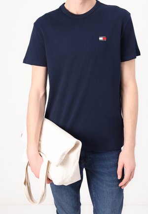 Personne portant un t-shirt Tommy Jeans bleu marine et un jean bleu, tenant un sac fourre-tout beige clair plié dans sa main gauche.