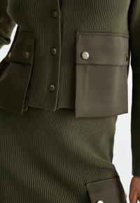 Cardigan a coste verde oliva con due ampie tasche a pattina, ciascuna dotata di bottoni a pressione argentati. Tessuto testurizzato e design aderente.