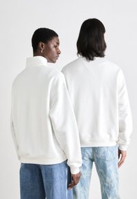 Sudadera de forro polar blanca con cuello alto, corte relajado, hombros caídos y dobladillo acanalado, combinada con jeans de patrón azul claro.