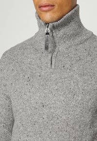 Homme portant un pull gris chiné en maille avec un col côtelé haut et une fermeture éclair argentée partiellement zippée avec une tirette en cuir.