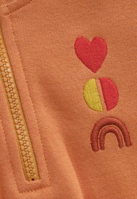 Orangenes Material mit einem gelben Reißverschluss und einem gestickten roten Herz, einem halb gelben und halb roten Kreis sowie einem roten Halbkreis-Regenbogen-Design.