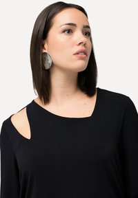 Schwarzes, leichtes Oberteil mit quadratischem Ausschnitt und Schulterauschnitten. Kombiniert mit großen, reflektierenden ovalen Ohrringen.