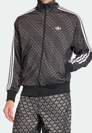 Survêtement Adidas noir et gris avec motif répétitif du logo trèfle, bandes blanches sur les manches, fermeture éclair à l’avant, poignets et ceinture côtelés.