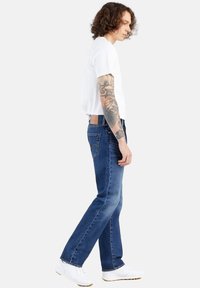 Jeans de denim azul con un corte ajustado, textura desgastada y bolsillos traseros. Combinados con una camiseta blanca básica y zapatillas blancas.
