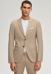 Mann trägt einen beigen Anzug mit zwei Knöpfen und Klappentaschen sowie ein weißes T-Shirt mit Rundhalsausschnitt, steht vor einem schlichten hellen Hintergrund.