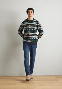 Gebreid sweater in donkergroen met gestreepte patronen in wit en goud. Gepaard met blauwe jeans en lichtbeige schoenen, staande op een houten vloer.