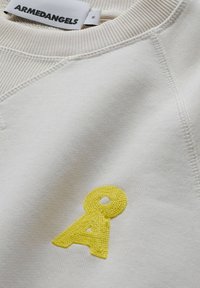 Cream-colored sweatshirt featuring a yellow embroidered "A" logo, textured fabric, and ribbed neckline with visible stitching details.
