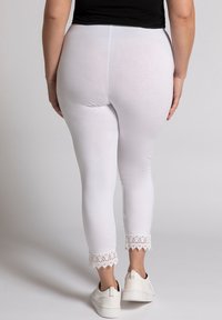 Person wearing white cropped leggings with lace trim at the ankles and white sneakers, shown from the back against a plain background.