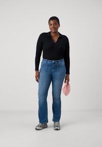 Musta pitkähiha bodysuit, jossa on polo-kaulus, yhdistettynä vaaleansinisiin suoriin farkkuihin. Pitelee vaaleanpunaista asustetta ja jalassa harmaat tennarit.