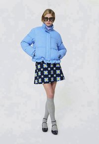 Kobieta w oversize'owej, jasnoniebieskiej kurtce puchowej, granatowej spódnicy w niebieskie i żółte kwiaty, szarych skarpetach do kolan, czarnych butach Mary Jane i okularach przeciwsłonecznych.