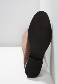 Brązowy zamszowy but z teksturowaną czarną gumową podeszwą. Posiada boczny zamek błyskawiczny i subtelne przeszycia na cholewce.