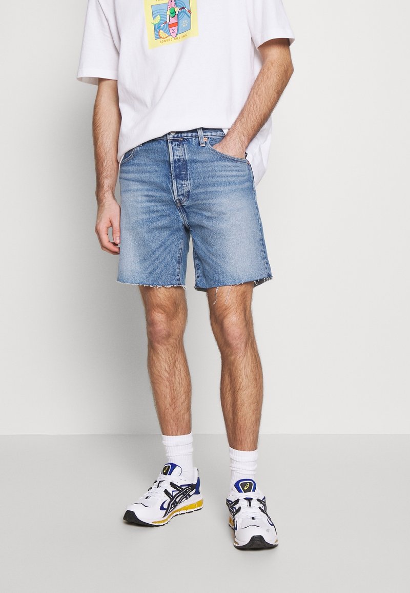 Shorts en denim bleu clair avec un ourlet effiloché, portés avec un t-shirt graphique blanc. Assortis à des chaussures de sport blanches et des chaussettes à côtes.