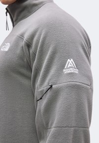 Κοντινό πλάνο ενός ατόμου που φοράει γκρι fleece μπουφάν με το λογότυπο "Mountain Athletics" στον πάνω βραχίονα και μια μικρή τσέπη με φερμουάρ από κάτω.