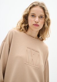 Beige sweatshirt med en strukturerad logga, har rund halsringning och sänkta axlar, tillverkad av ett mjukt tyg med en slät yta.