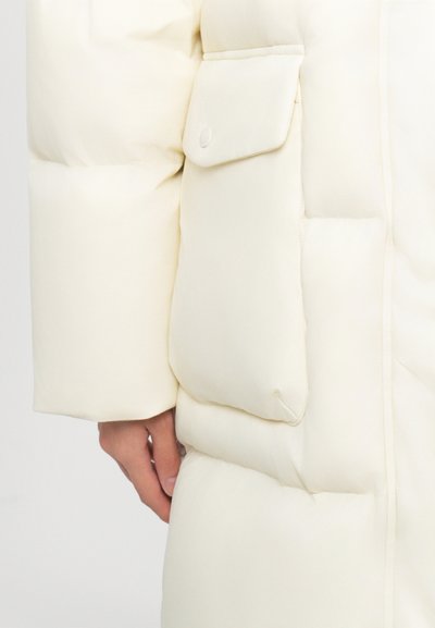 Beige Puffer-Jacke mit einer großen Seitentasche und Knopf, strukturiertem Stoff und gestepptem Muster an den Ärmeln.