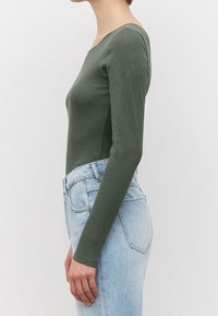 Langärmliger Body aus olivgrünem Rippmaterial. Mit einem runden Ausschnitt und einer figurbetonten Silhouette, kombiniert mit hellblauen High-Waist-Jeans.