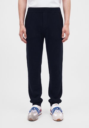 Homme portant un pantalon de survêtement noir avec revers, associé à des baskets blanches avec des lacets bleus et une chemise blanche.