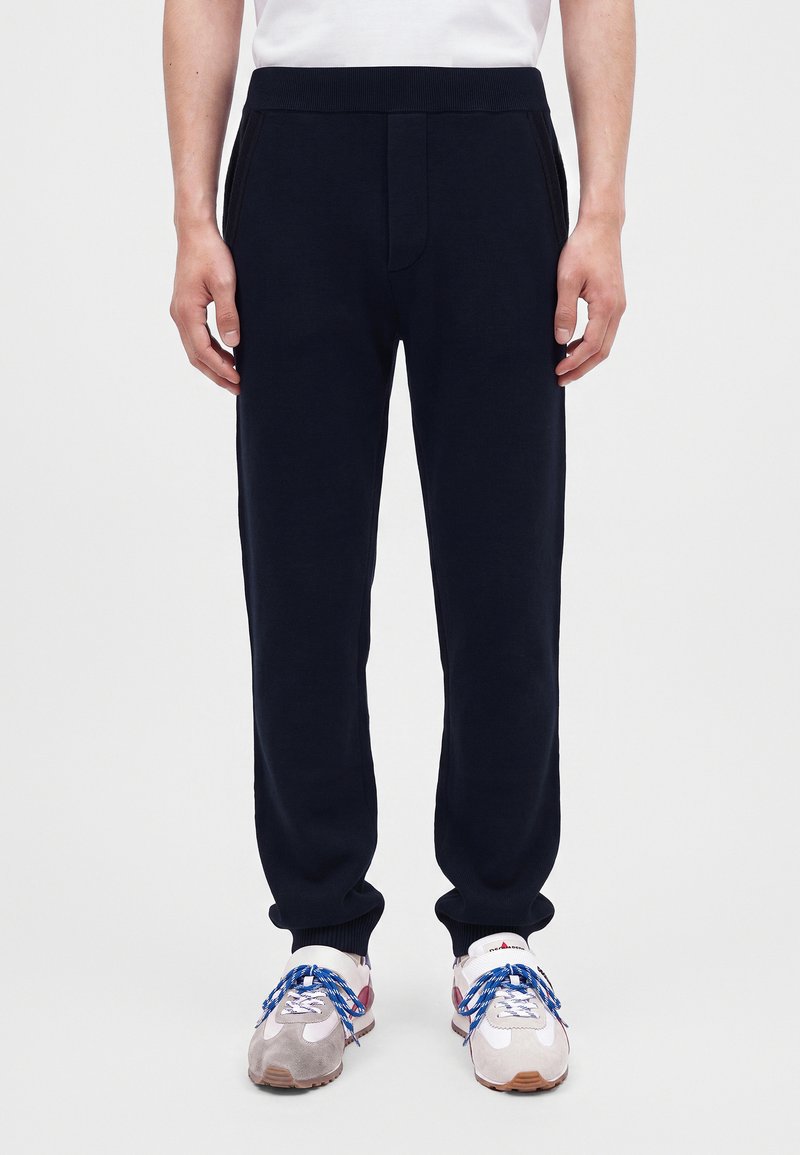 Homme portant un pantalon de survêtement noir avec revers, associé à des baskets blanches avec des lacets bleus et une chemise blanche.