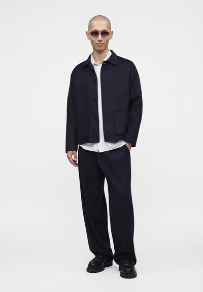 Homme portant des lunettes de soleil, une veste sombre avec de grandes poches, une chemise blanche, un pantalon sombre et des chaussures noires, debout devant un fond blanc uni.