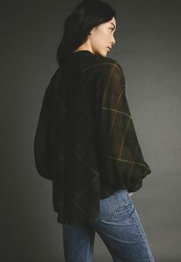 Blouse noire transparente avec un motif à carreaux jaune et rouge, coupe décontractée, manches longues. Associée à un jean bleu taille mi-haute. Tissu texturé visible.