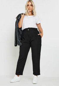 Pantalones negros ajustados con cintura alta, pliegues, bolsillos laterales y puños enrollados, combinados con una camiseta blanca y una chaqueta de cuero negra.