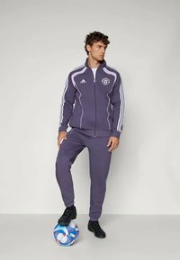 Junger Mann in einem lila Adidas-Trainingsanzug mit Manchester United-Logos steht mit einem Fuß auf einem blau-weißen Fußball vor einem einfarbigen Hintergrund.