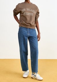 Brown sweatshirt with "New Balance Athletics" text, blue straight-leg jeans, and white sneakers with blue accents. Model stands on a yellow background.
