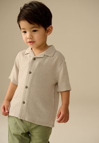 Lichtbeige shirt met korte mouwen en een kraag, voorzien van bruine knopen en een gestructureerde stof. Gecombineerd met groene broek.