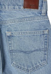 Svetlo modra denim tkanina z zadnjim žepom. Opremljena s temno rjavim usnjenim oznako z vtisnjenim logotipom. Vidna podrobnost šivanja okoli žepa.