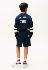 Junge, der sich wegdreht und navyblaue Shorts sowie ein Sweatshirt mit "CLASS OF 1985" auf der Rückseite trägt, weiße Sneakers und rotgestreifte Socken.